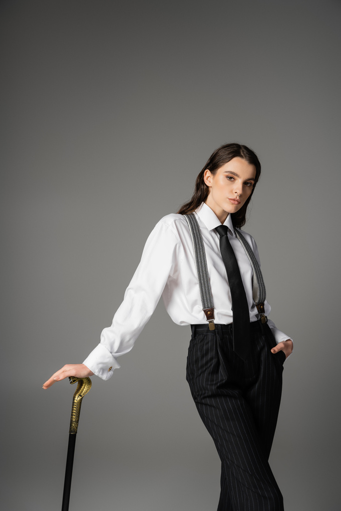pretty brunette model in white shirt and tie posing with walking cane and hand in pocket of black pants isolated on grey - Foto, Imagem