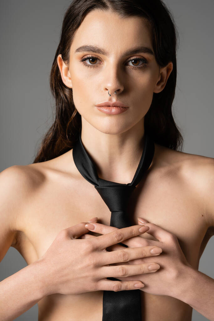 front view of shirtless woman in black tie covering breast with hands and looking at camera isolated on grey - Fotografie, Obrázek
