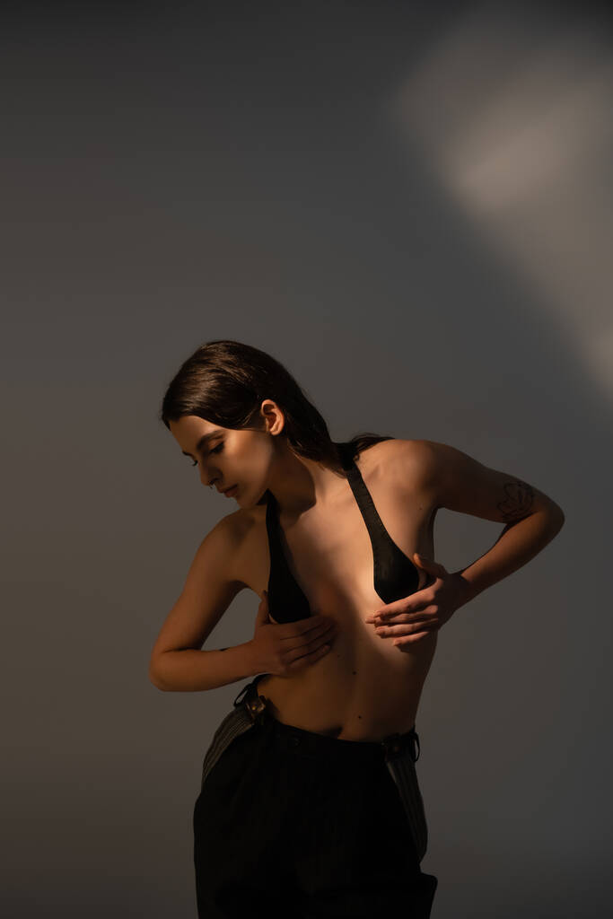 брюнетка женщина без рубашки тело позирует в черных брюках и грудной ленты на сером фоне с освещением - Фото, изображение