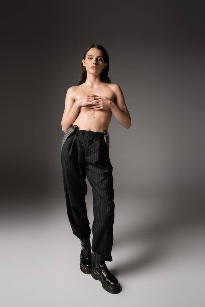 shirtless woman with wet body covering breast with hands while posing in black pants and rough boots on grey  - Foto, Bild