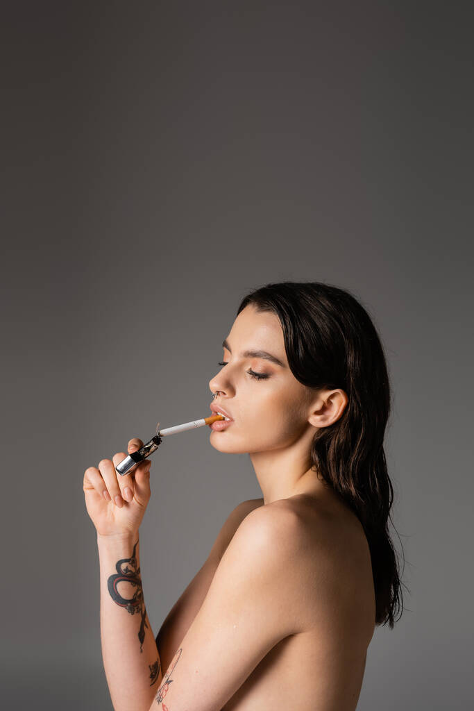 боковой вид на татуированную женщину без рубашки, закуривающую сигарету, изолированную на сером - Фото, изображение