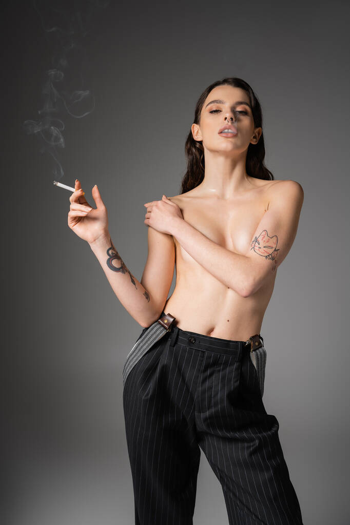 young shirtless woman in black striped pants covering bust and looking at camera while smoking on grey background - Zdjęcie, obraz