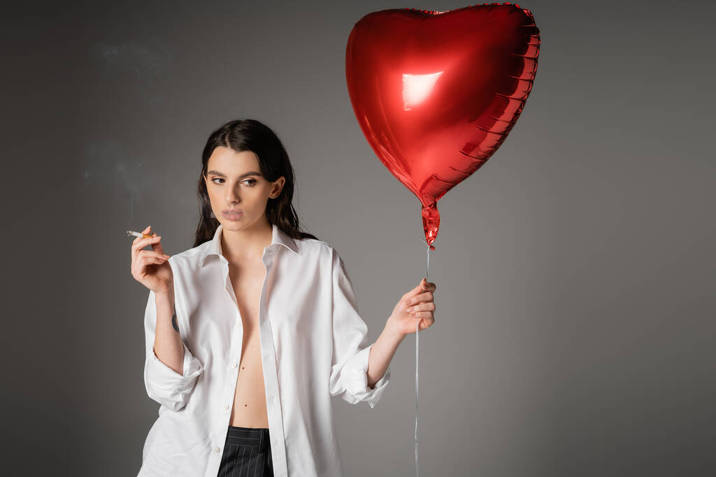 sexy woman in white unbuttoned shirt posing with cigarette and red heart-shaped balloon on grey background - Fotoğraf, Görsel