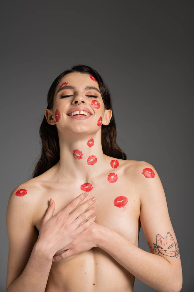 joyful shirtless woman with red kisses on face and body covering breast with hands isolated on grey - Φωτογραφία, εικόνα