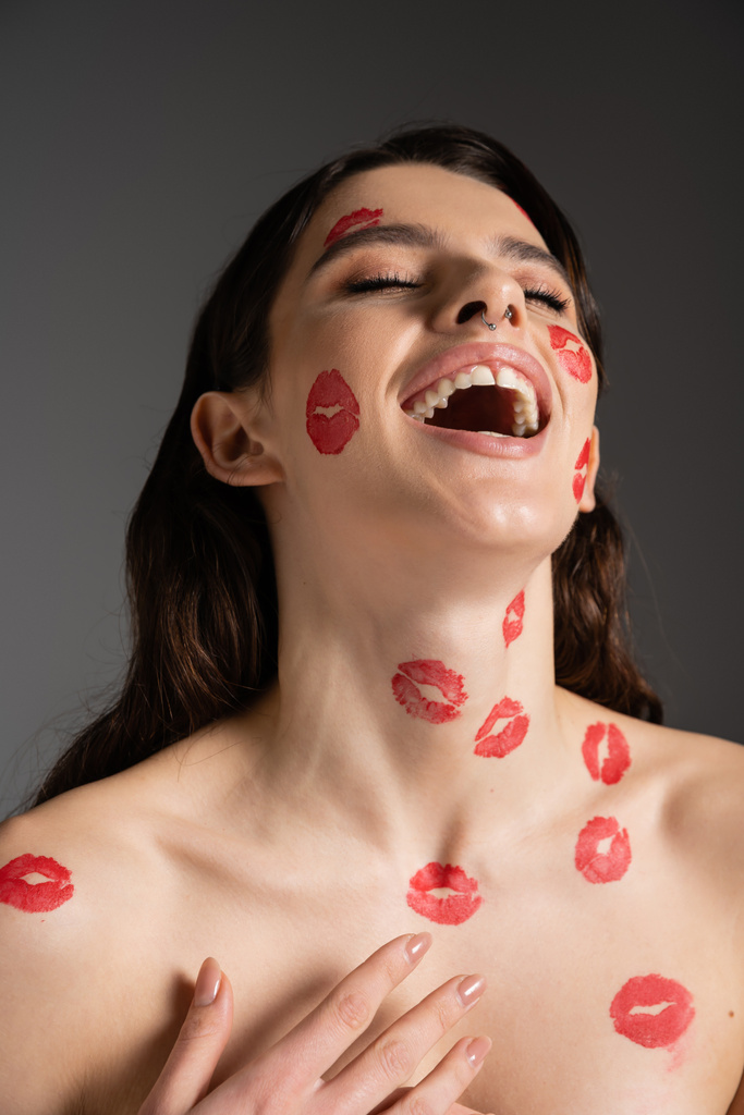 excited woman with closed eyes and red kisses on face and naked shoulders laughing isolated on grey - Photo, Image