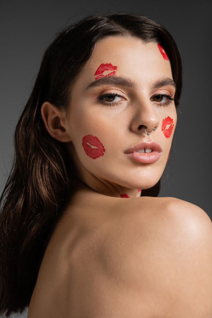 portrait of brunette woman with bare shoulder and red kiss prints looking at camera isolated on grey - Fotó, kép