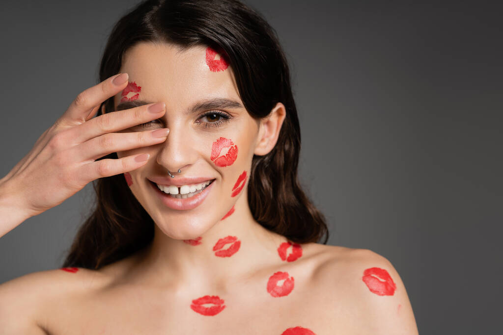 sexy brunette woman with red lipstick marks on bare shoulders and face covering eye with hand and smiling isolated on grey - Zdjęcie, obraz