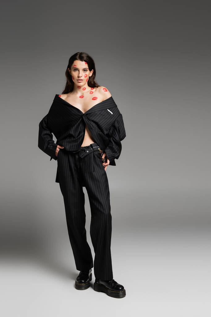 full length of brunette woman with red kiss prints wearing black oversize suit and looking at camera on grey  - Fotoğraf, Görsel