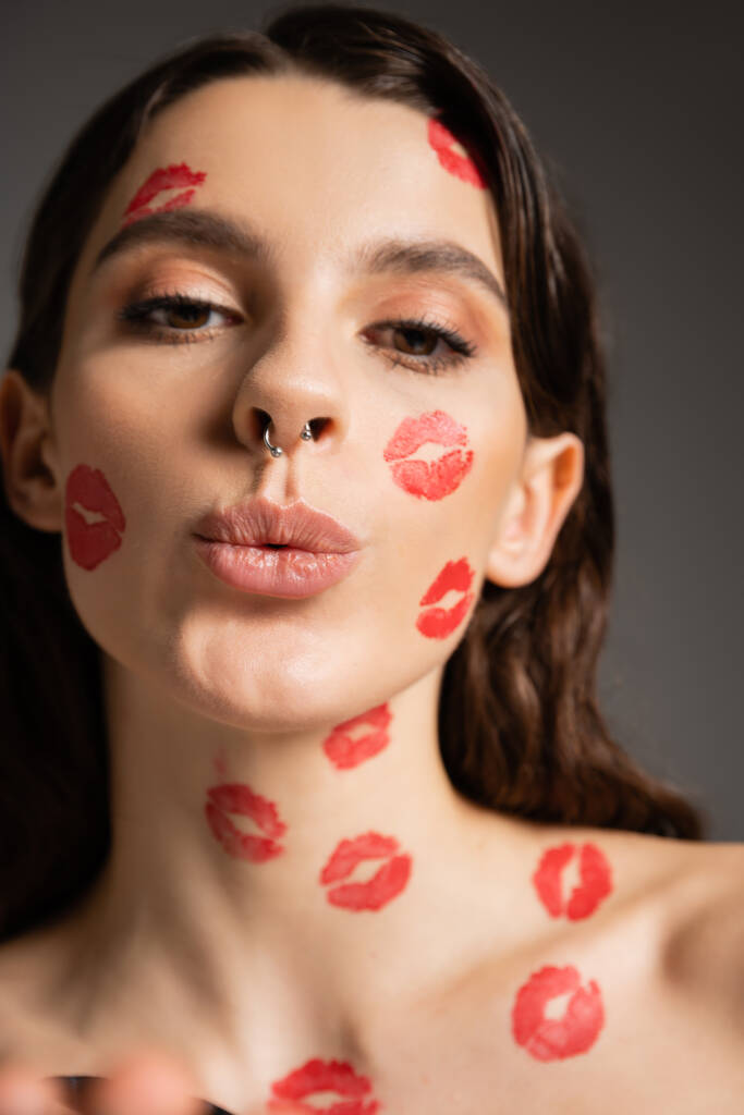 portrait of pretty woman with makeup and red kiss prints pouting lips while looking at camera isolated on grey - Zdjęcie, obraz