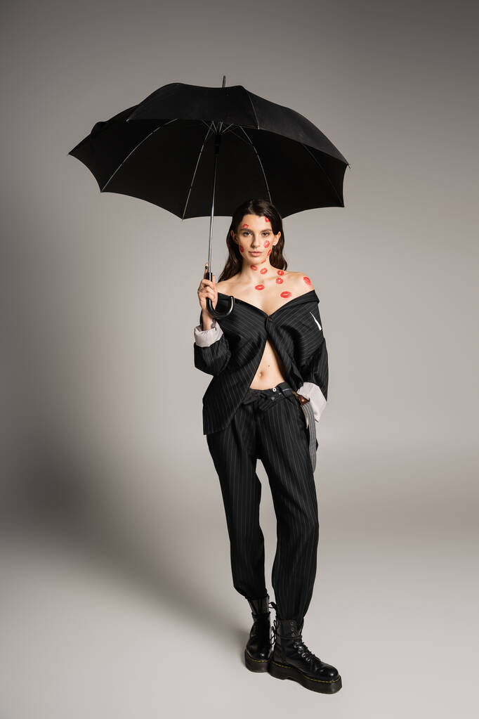 full length of sexy woman with red kiss prints wearing oversize suit while holding umbrella on grey background - Foto, afbeelding