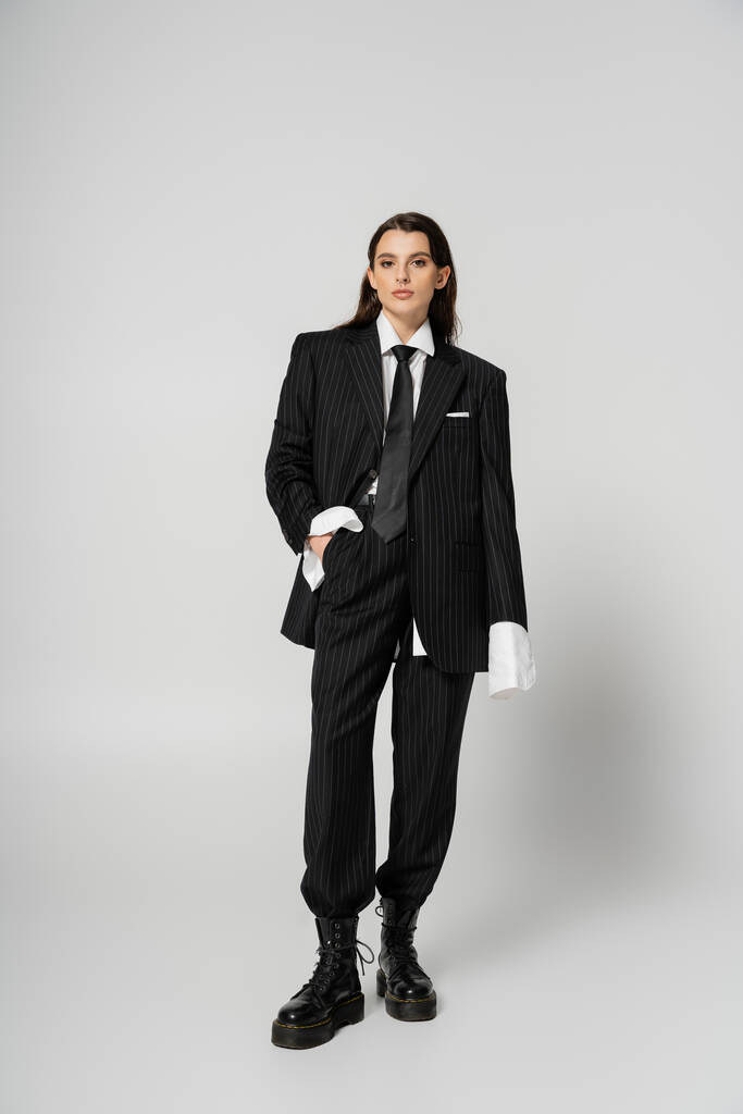 Полная длина брюнетки женщина в стильной формальной одежде и грубой кожаные сапоги позируя с рукой в кармане на сером  - Фото, изображение