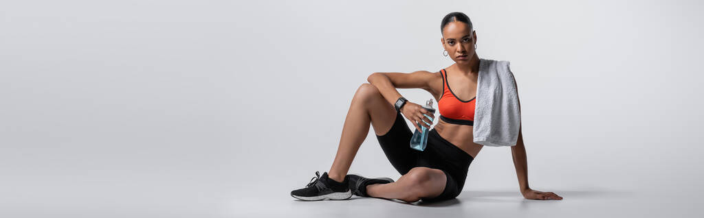 young african american woman in sports bra and bike shorts sitting with sports bottle and towel on grey background, banner  - Valokuva, kuva