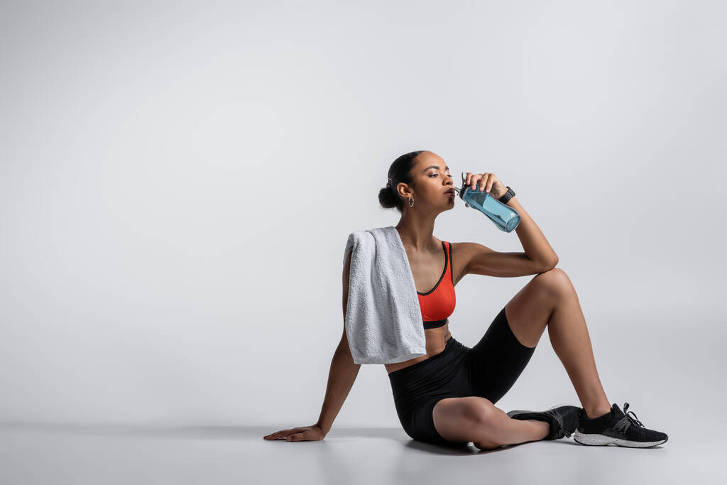 young african american woman in sports bra and bike shorts sitting with towel and drinking water on grey background  - Fotó, kép