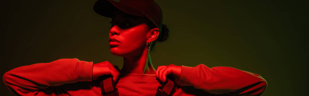african american woman in sweatshirt and baseball cap posing on green with red light, banner  - 写真・画像