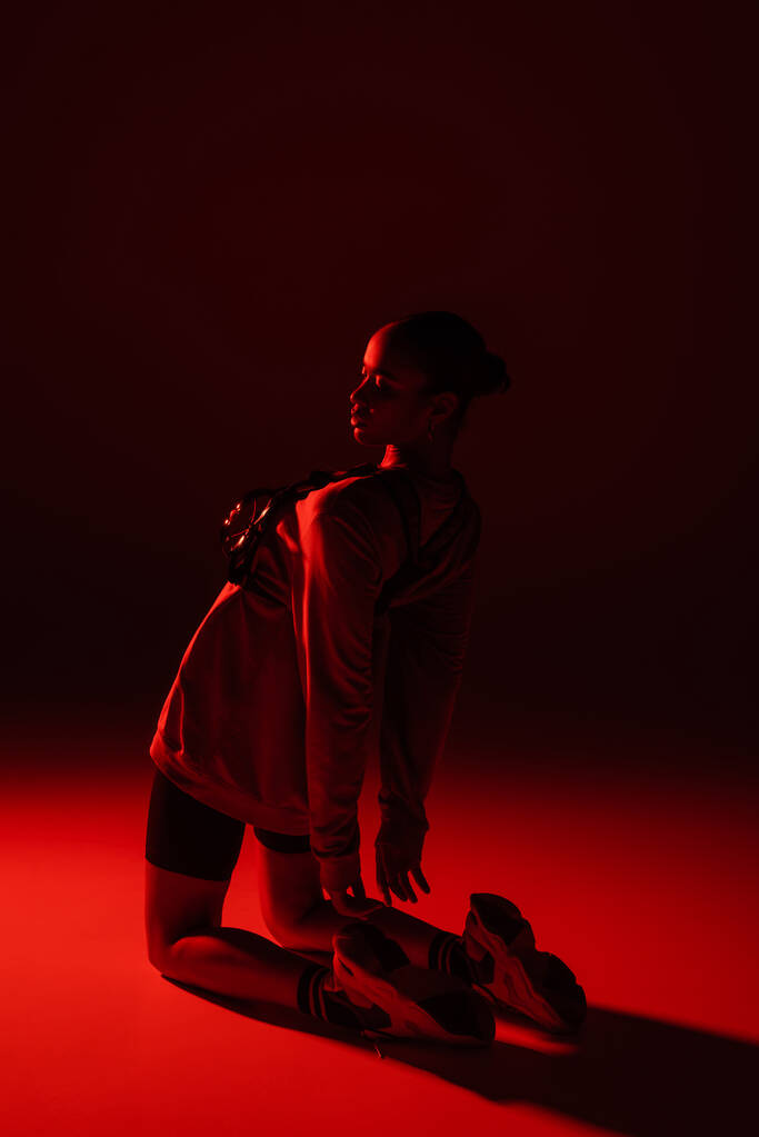full length of young african american woman in bike shorts and sweatshirt posing on dark red background  - Foto, Imagem