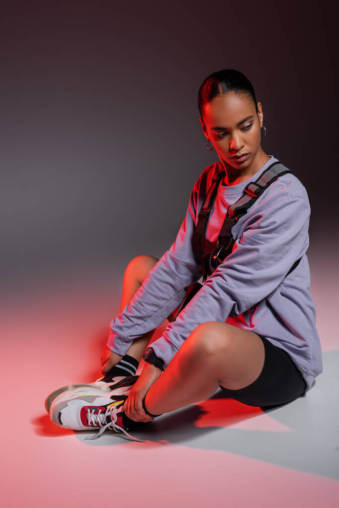 full length of stylish african american woman in bike shorts and sweatshirt sitting on grey with red light  - Photo, Image