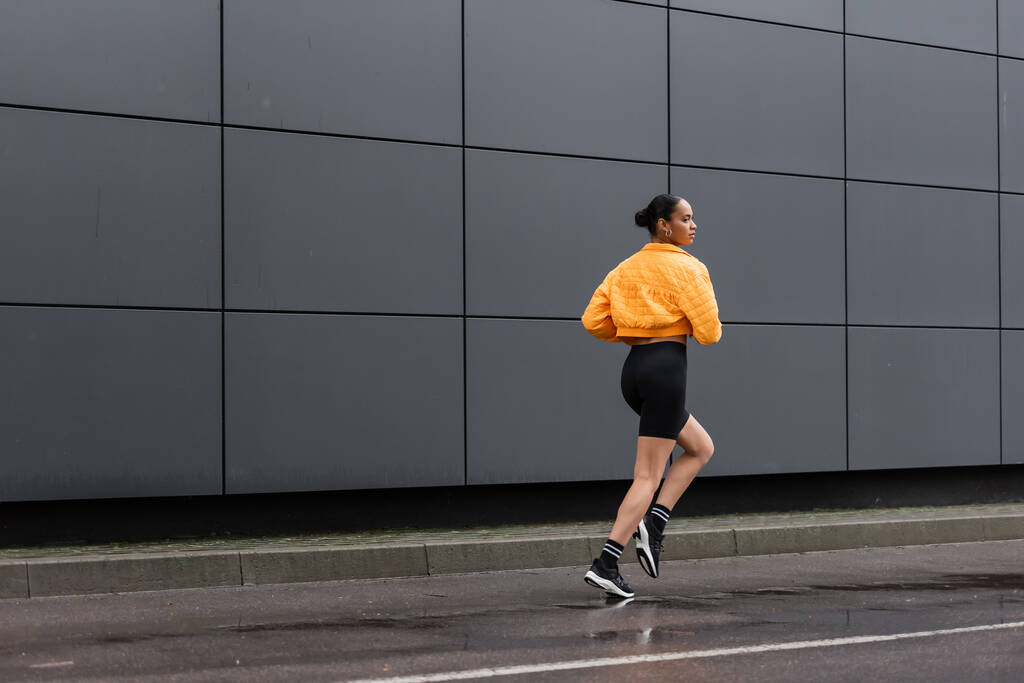 full length of brunette african american sportswoman in bike shorts and yellow puffer jacket jogging outside  - Photo, Image