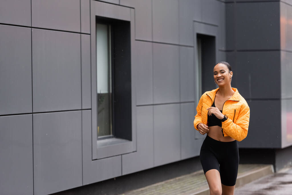 positive african american sportswoman in bike shorts and yellow puffer jacket jogging  outside  - Φωτογραφία, εικόνα