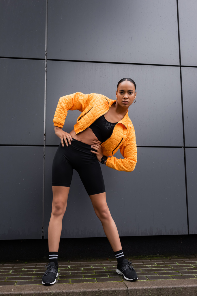 full length of brunette african american sportswoman in bike shorts and yellow puffer jacket exercising with hands on hips  - Fotografie, Obrázek