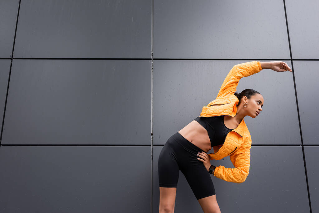 brunette african american sportswoman in bike shorts and yellow puffer jacket exercising with hand on hip  - Foto, afbeelding