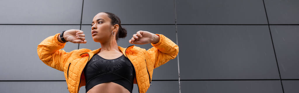 low angle view of brunette african american sportswoman in sports bra and yellow puffer jacket exercising outside, banner  - Foto, Imagem