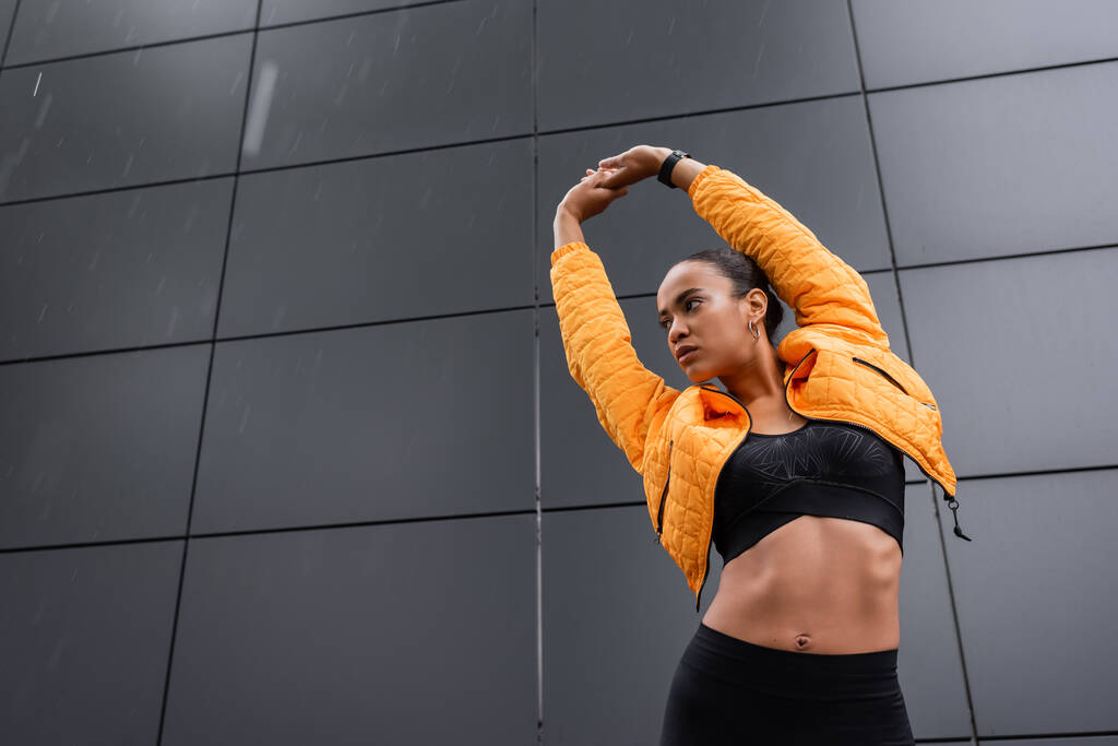 low angle view of african american sportswoman in sports bra and yellow puffer jacket warming up outside  - Foto, afbeelding