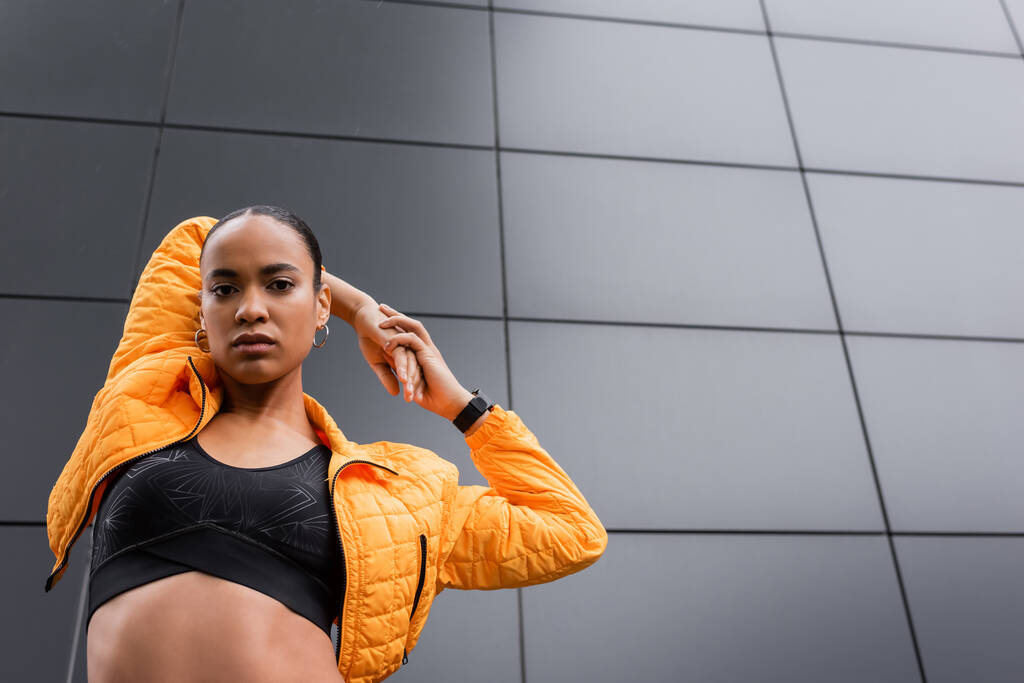 low angle view of african american sportswoman in sports bra and puffer jacket warming up near grey building  - 写真・画像