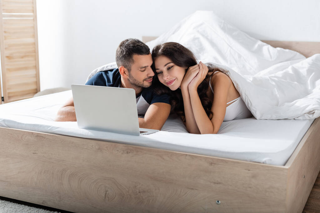 Бородатый мужчина смотрит на девушку возле ноутбука на кровати дома  - Фото, изображение
