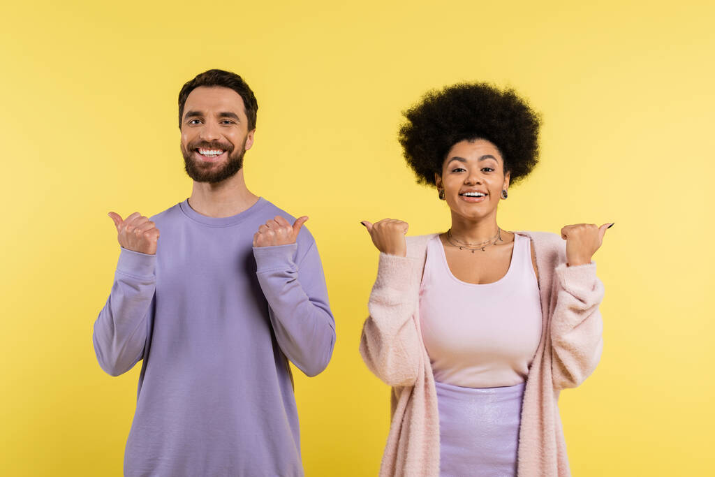 cheerful interracial couple pointing with thumbs and looking at camera isolated on yellow - Foto, immagini