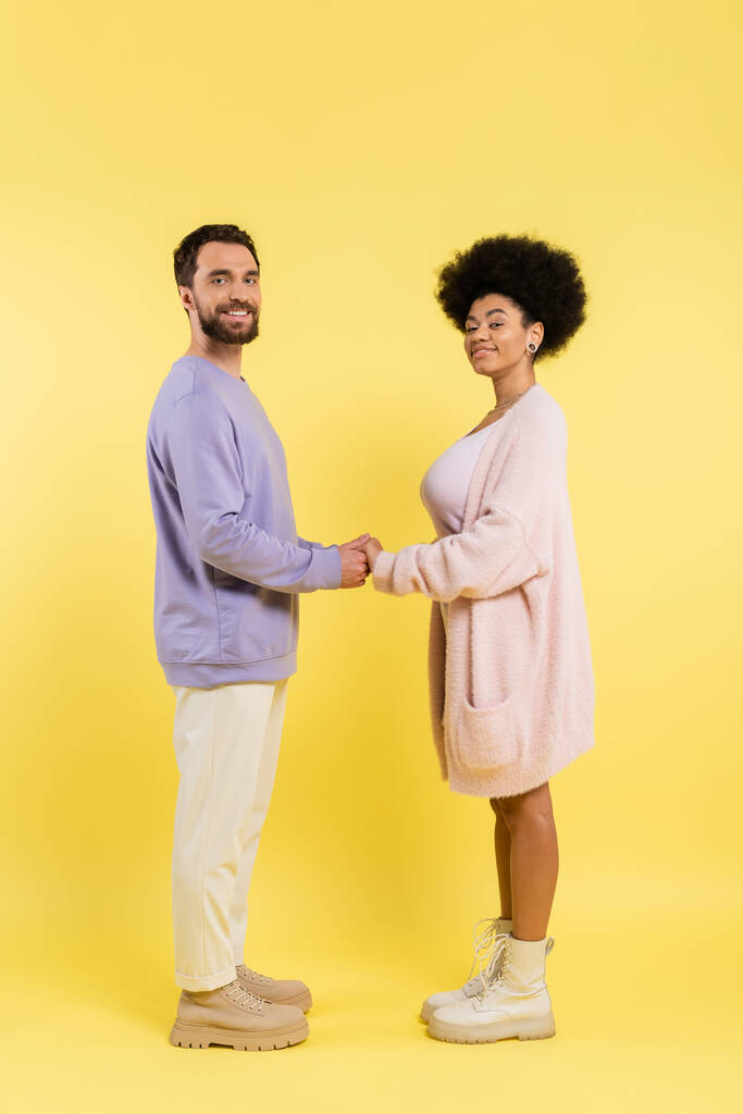 full length of interracial couple in stylish clothes holding hands and looking at camera on yellow background - Fotó, kép