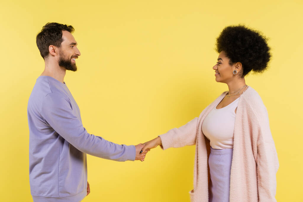 side view of bearded man and curly african american woman shaking hands and smiling at each other isolated on yellow - Photo, image
