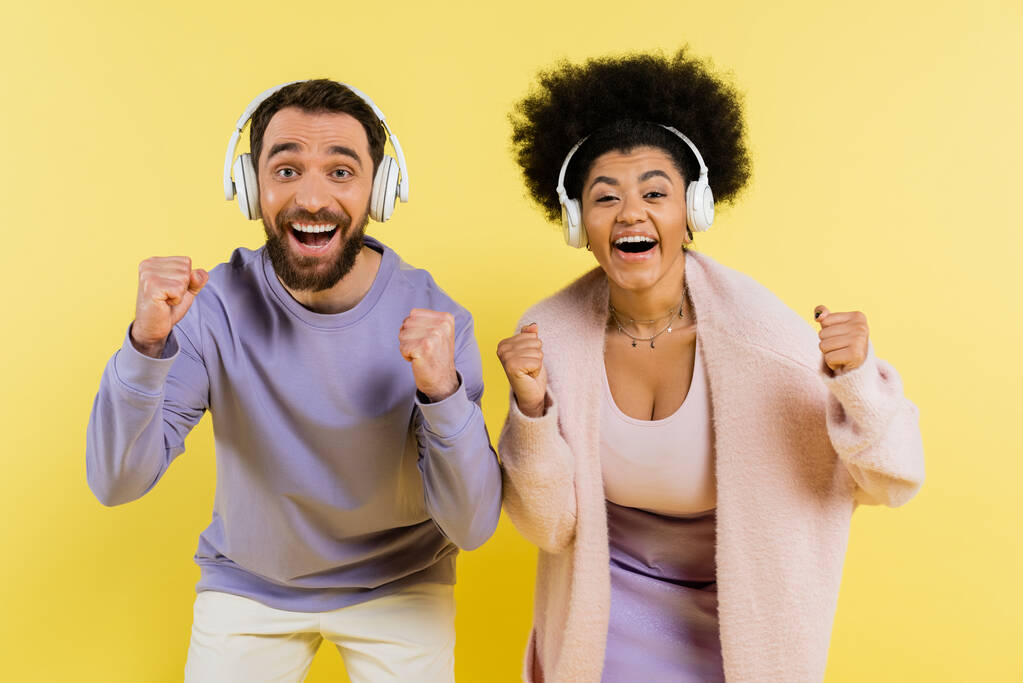 excited interracial couple in wireless headphones showing yeah gesture isolated on yellow - Φωτογραφία, εικόνα