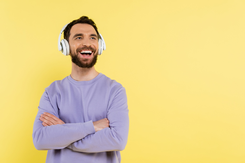 無線のヘッドフォンで音楽を聴いている髭の男が腕を組んで立って黄色で孤立して笑っている - 写真・画像