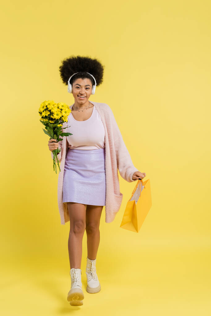 full length of smiling african american woman in wireless headphones walking with flowers and shopping bag on yellow background - Fotoğraf, Görsel