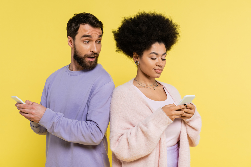 Любопытный мужчина с открытым ртом, глядя на африканскую американку, обменивающуюся сообщениями на смартфоне, изолированном на желтый - Фото, изображение