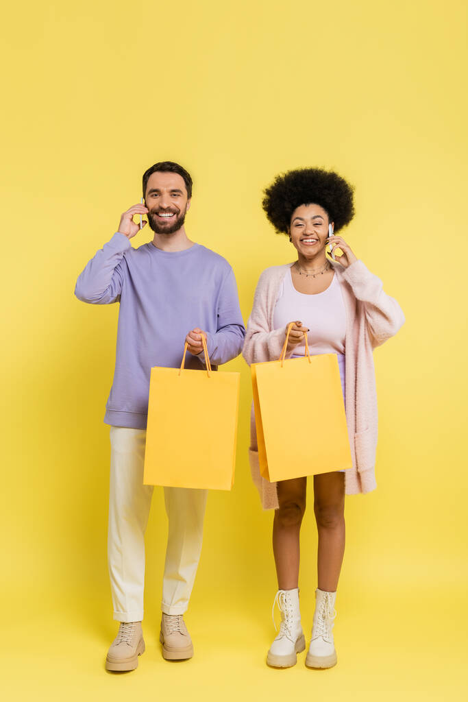 full length of happy interracial couple holding shopping bags and talking on smartphones on yellow background - Fotoğraf, Görsel