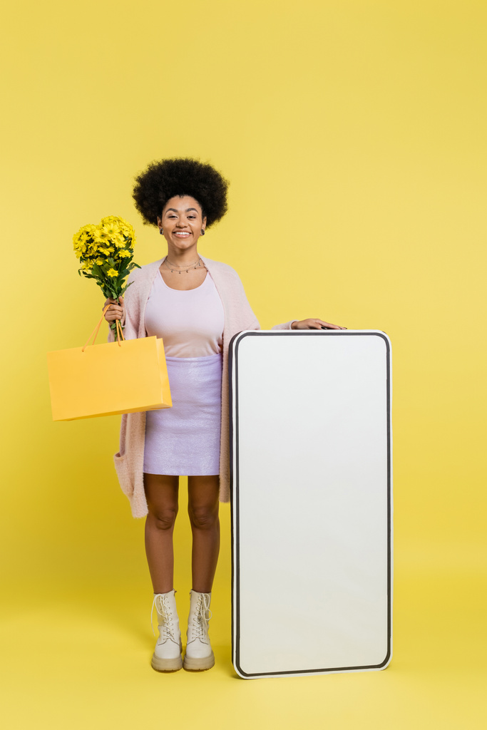 full length of joyful african american woman with flowers and shopping bag near white template of mobile phone on yellow background - Foto, afbeelding