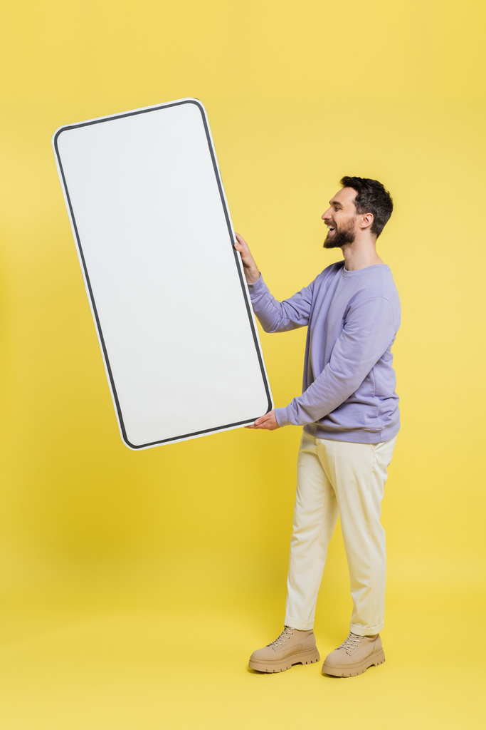 side view of happy bearded man holding huge template of mobile phone on grey background - Photo, image
