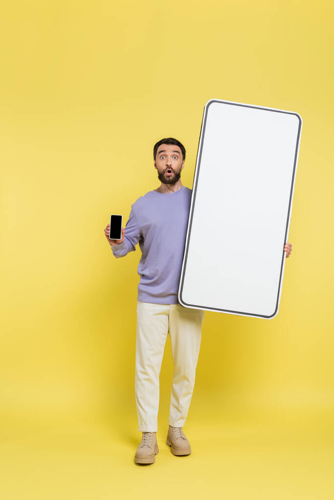 полная длина удивленный человек показывает смартфон с пустым экраном рядом с белым шаблоном мобильного телефона на сером фоне - Фото, изображение