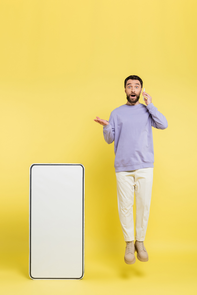amazed man talking on cellphone while levitating and pointing at huge mock-up of mobile phone on grey background - Foto, Imagen