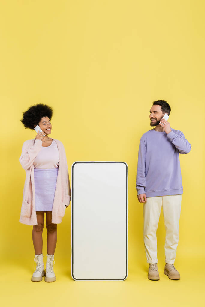 full length of interracial couple talking on cellphone and smiling at each other near blank mock-up of smartphone on yellow  - Foto, immagini