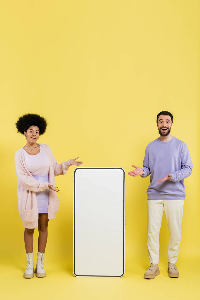 full length of pleased multiethnic couple looking at camera and pointing at huge mobile phone template on yellow background - Fotoğraf, Görsel