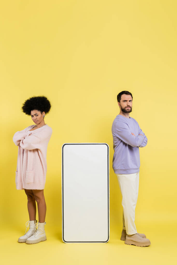 full length of displeased interracial couple standing with crossed arms near huge phone template on yellow background - Foto, Imagen