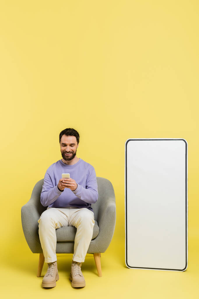 full length of pleased man using smartphone while sitting in armchair near huge template of mobile phone on grey background - Foto, Bild