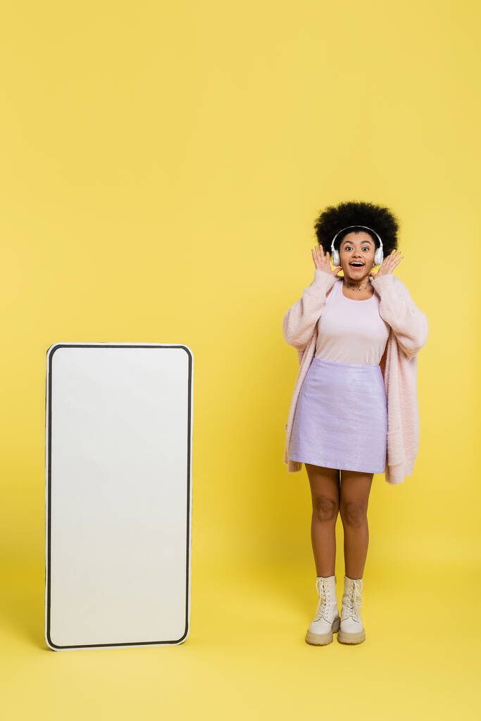 full length of excited african american woman in wireless headphones showing wow gesture near huge template of smartphone on yellow  - Zdjęcie, obraz