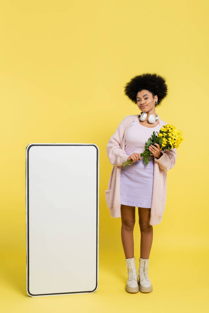 full length of african american woman with wireless headphones and flowers smiling near blank template of smartphone on yellow  - Photo, image