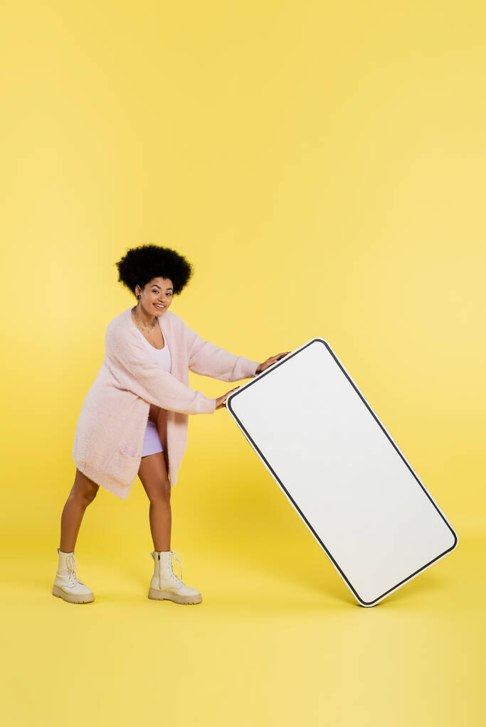 full length of smiling african american woman in cardigan and boots holding carton template of cellphone on yellow background - Foto, Imagem