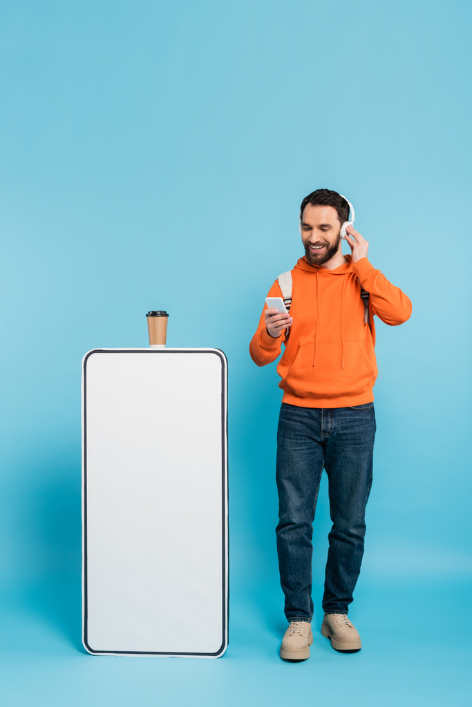 full length of bearded student in wireless headphones standing with smartphone near phone mock-up and paper cup on blue background - Fotó, kép