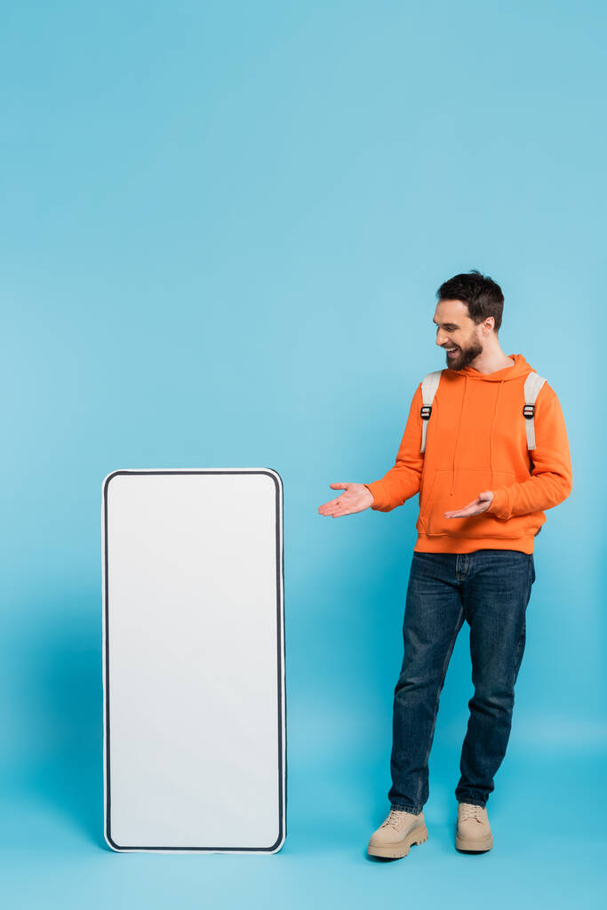 full length of happy bearded student pointing at big template of mobile phone on blue background - Photo, Image