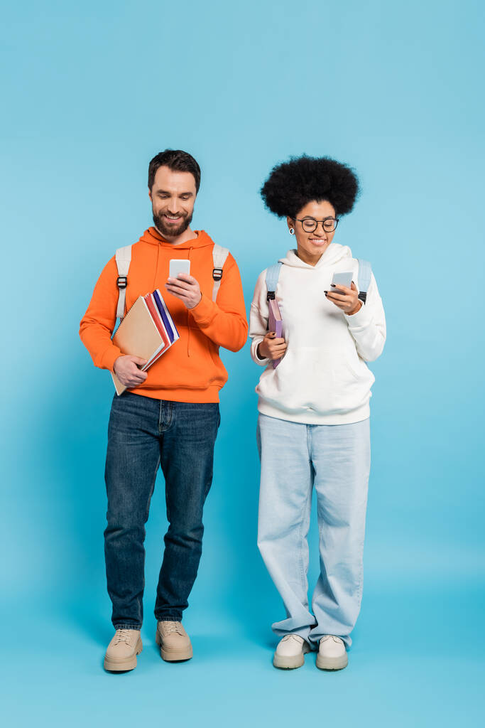 полная длина счастливых мультиэтнических студентов с рюкзаками и ноутбуками с использованием смартфонов на голубом  - Фото, изображение